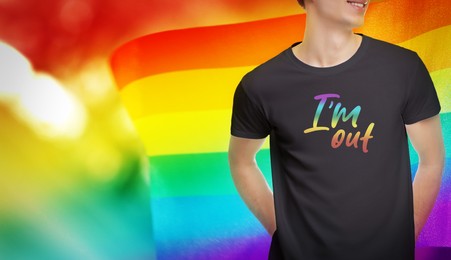 Image of Coming out. Man wearing black t-shirt with phrase I'm Out in rainbow colors against pride flag, closeup