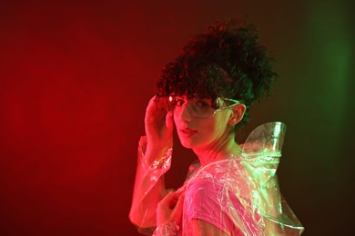 Beautiful young woman in transparent coat and sunglasses posing on color background in neon lights