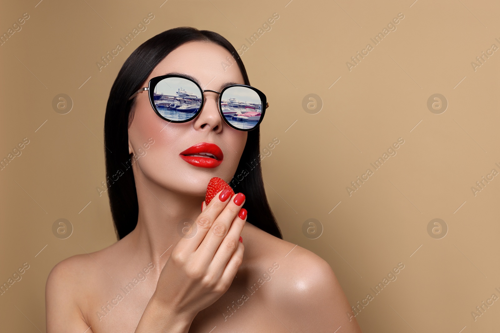 Image of Attractive woman in stylish sunglasses holding strawberry on dark beige background. Yachts reflecting in lenses. Space for text