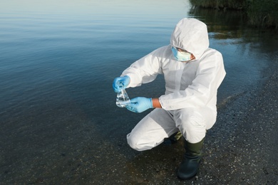 Scientist in chemical protective suit with conical flask taking sample from river for analysis