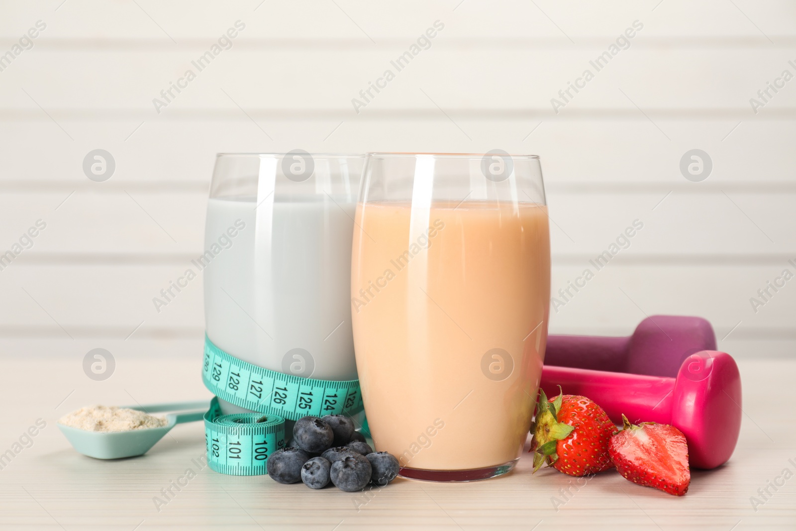 Photo of Tasty shakes with berries, dumbbells, measuring tape and powder on light wooden table. Weight loss