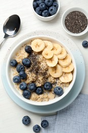 Tasty oatmeal with banana, blueberries and chia seeds served in bowl on white wooden table, flat lay