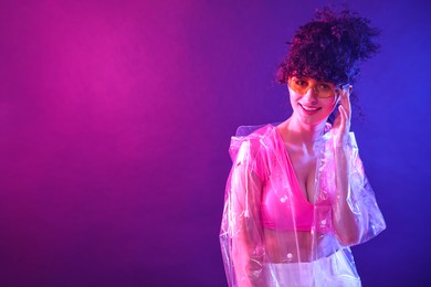 Photo of Beautiful young woman in transparent coat and sunglasses posing on color background in neon lights. Space for text