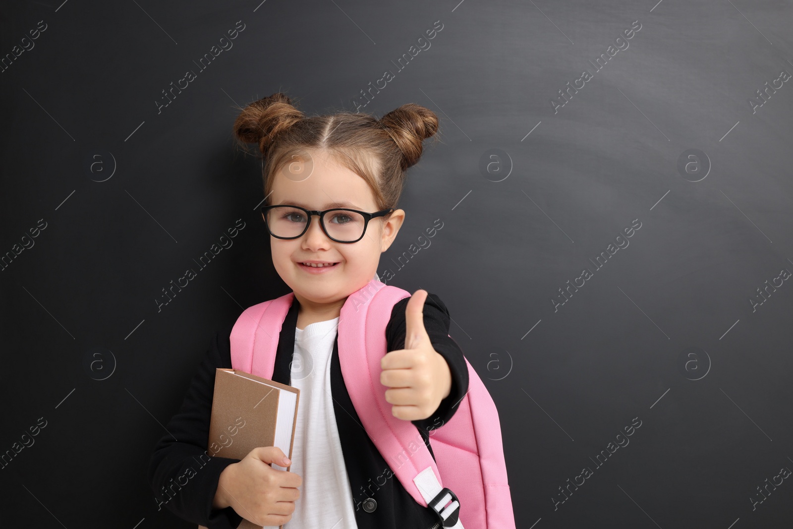 Photo of Happy little school child with notebook showing thumbs up near chalkboard. Space for text