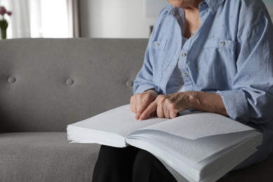 Blind senior person reading book written in Braille on sofa indoors, closeup. Space for text