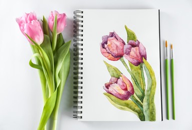 Photo of Painting of tulips in sketchbook, flowers and brushes on white background, flat lay