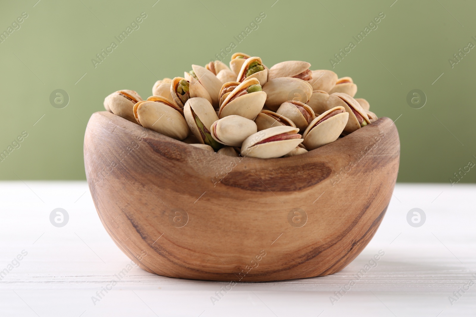 Photo of Tasty pistachios in bowl on white wooden table against olive background, closeup