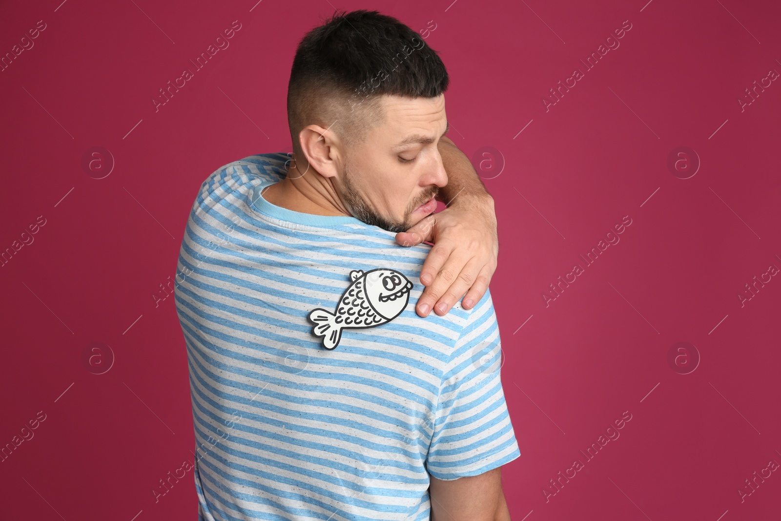 Photo of Man with paper fish on back against pink background. April fool's day