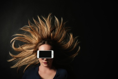 Young woman with smartphone covering her eyes on black background, top view. Space for text