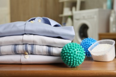 Photo of Dryer balls near stacked clean clothes and detergent on wooden table in laundry room, closeup
