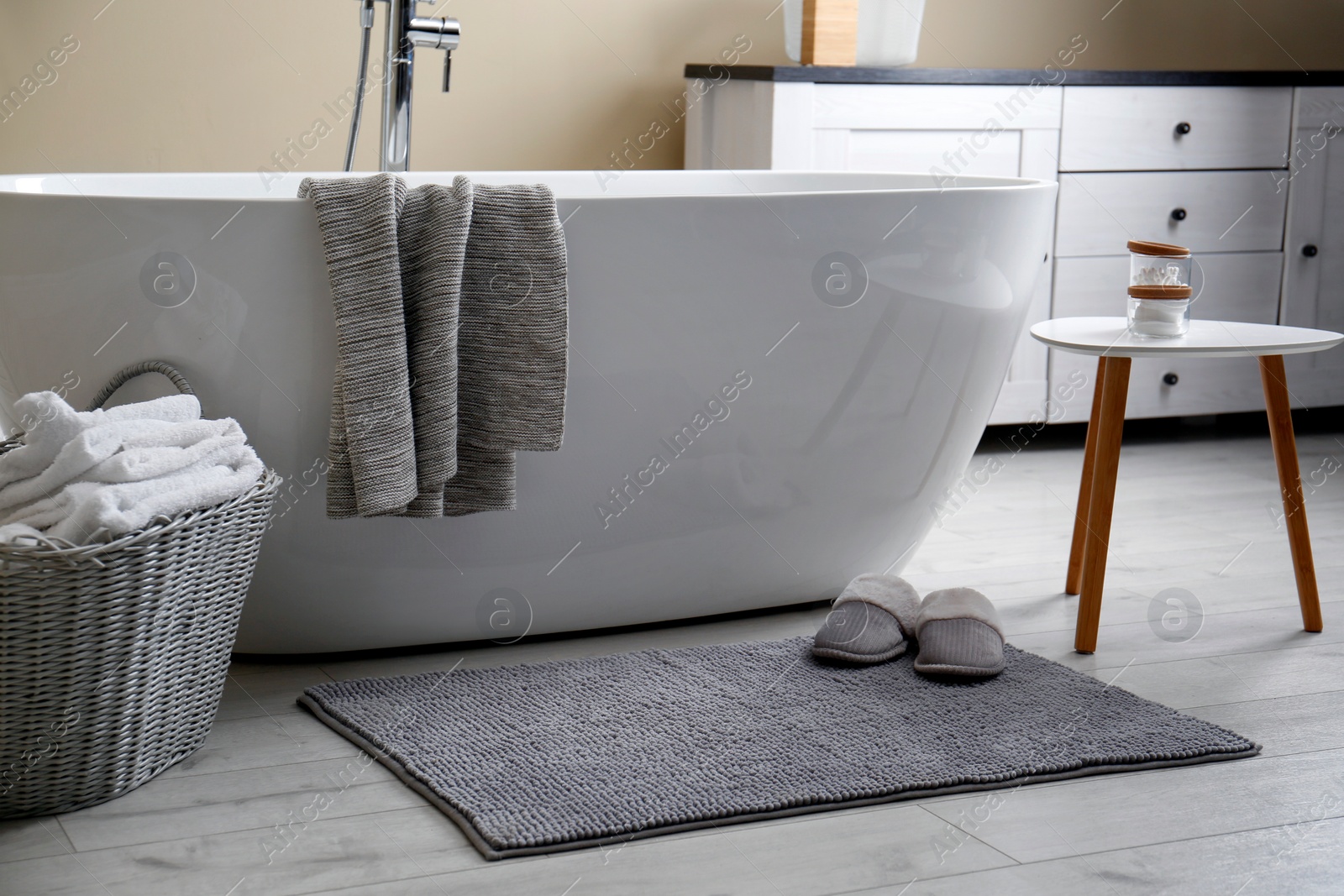 Photo of Soft grey mat with slippers on floor near tub in bathroom. Interior design