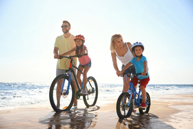 Happy parents teaching children to ride bicycles on sandy beach near sea