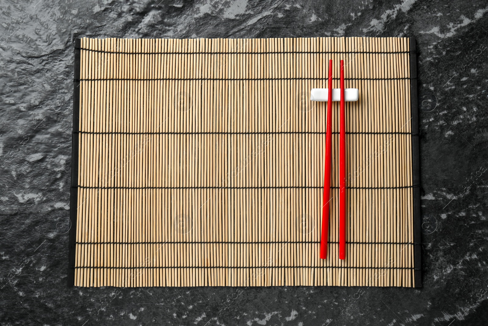 Photo of Bamboo mat with pair of red chopsticks and rest on black textured table, top view