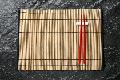 Bamboo mat with pair of red chopsticks and rest on black textured table, top view