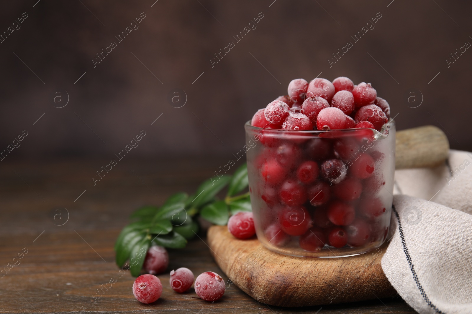 Photo of Frozen red cranberries in glass pot and green leaves on wooden table, closeup. Space for text