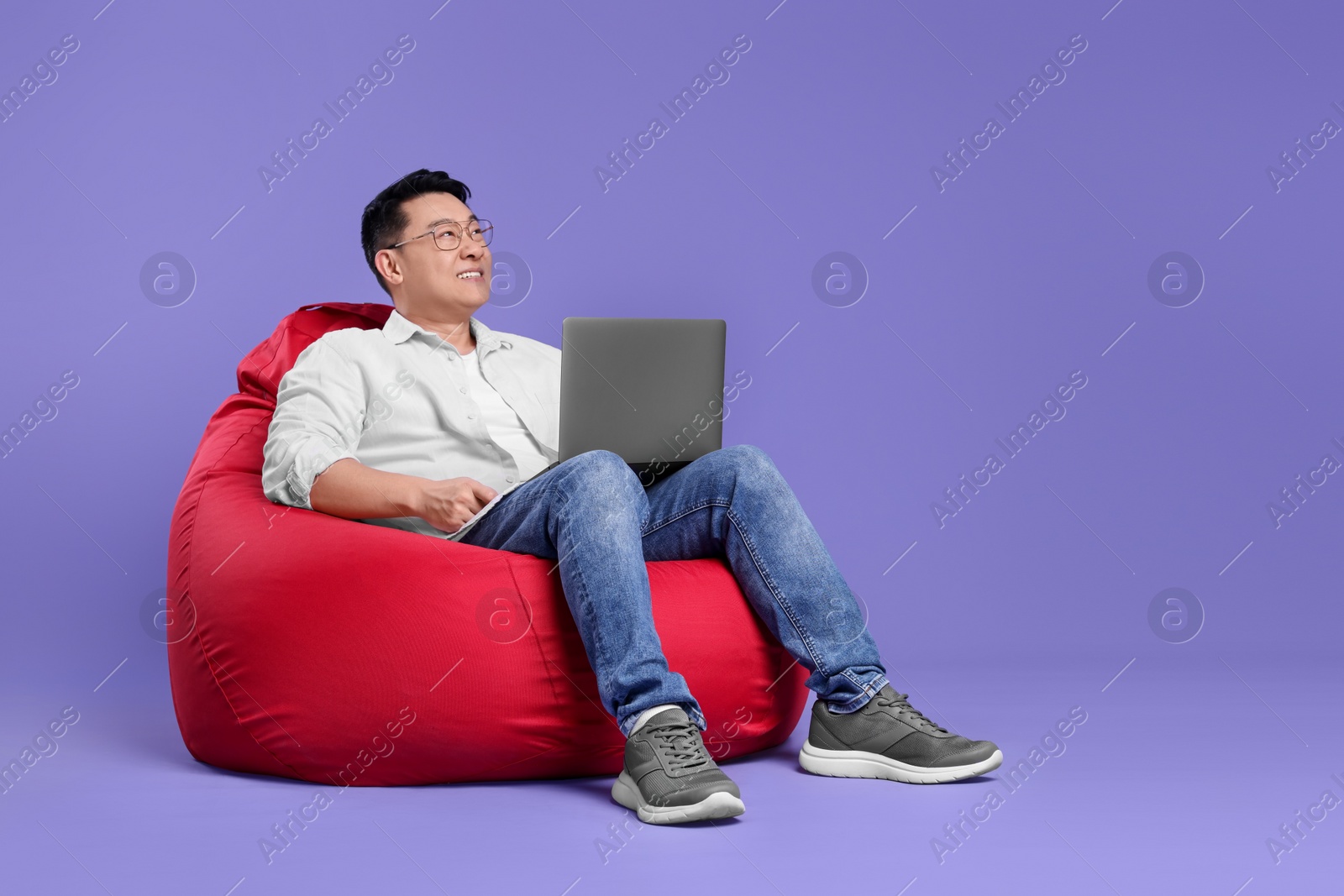 Photo of Smiling man with laptop sitting in beanbag chair against lilac background, space for text