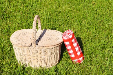 Photo of Rolled checkered tablecloth near picnic basket on green grass outdoors, space for text