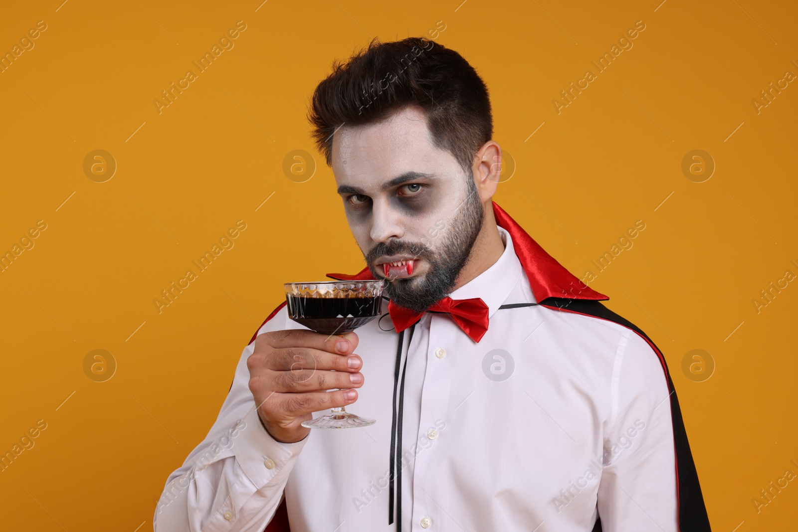 Photo of Man in scary vampire costume with fangs and glass of wine on orange background. Halloween celebration