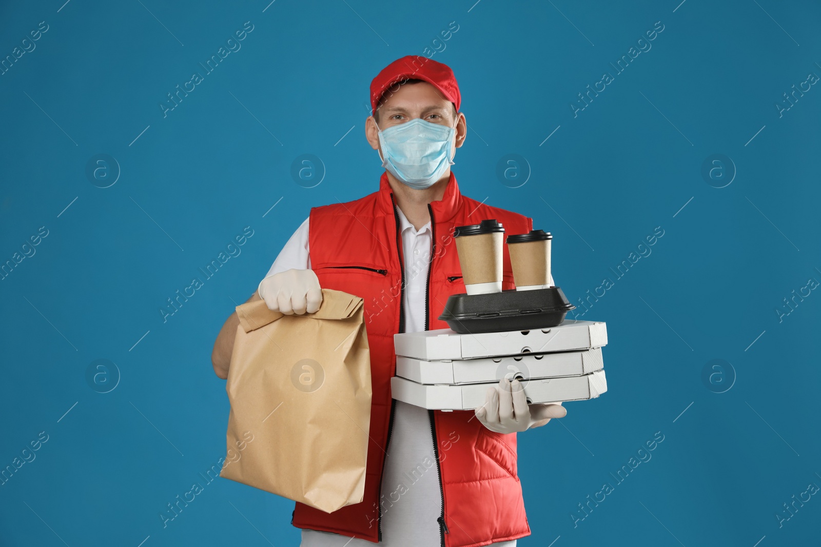 Photo of Courier in protective mask and gloves holding order on blue background. Food delivery service during coronavirus quarantine