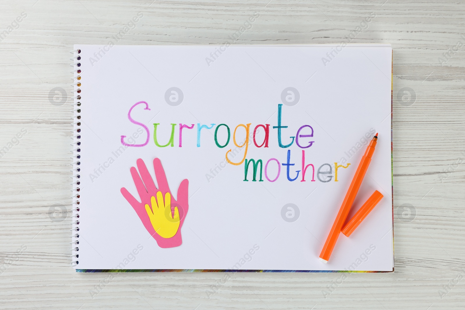 Photo of Sketchbook with phrase Surrogate mother, paper palms and felt tip pen on white wooden table, top view