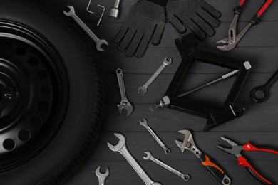 Car wheel, scissor jack, gloves and different tools on black wooden surface, flat lay
