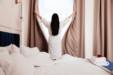 Photo of Young woman opening window curtains in hotel room, back view
