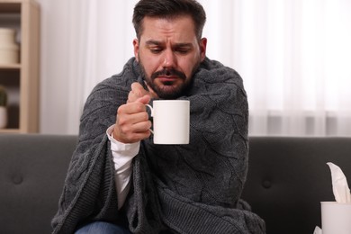 Photo of Sick man wrapped in blanket with cup of drink on sofa at home. Cold symptoms