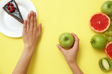 Photo of Top view of woman choosing between cake and healthy fruits on yellow background, closeup