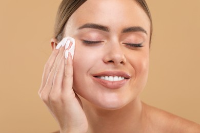 Beautiful woman removing makeup with cotton pad on beige background, closeup