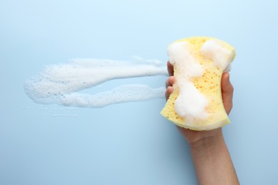 Man holding sponge with foam on light blue background, top view