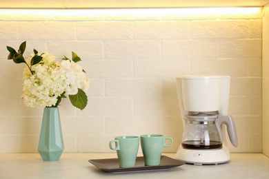 Bouquet with beautiful white hydrangea flowers, coffee maker and cups on light countertop