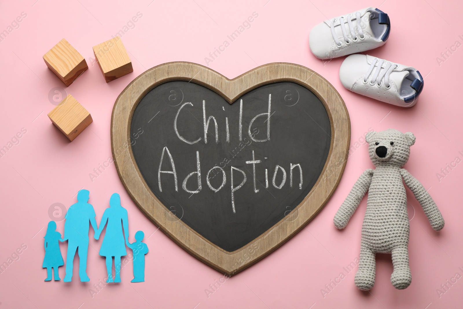 Photo of Heart shaped chalkboard with phrase Child Adoption, toy bear, baby shoes, paper family figure and wooden cubes on pink background, flat lay