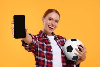 Photo of Emotional fan holding football ball and showing smartphone on yellow background, selective focus