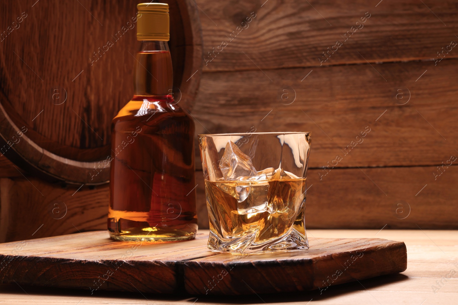 Photo of Whiskey with ice cubes in glass, bottle and barrel on wooden table, space for text