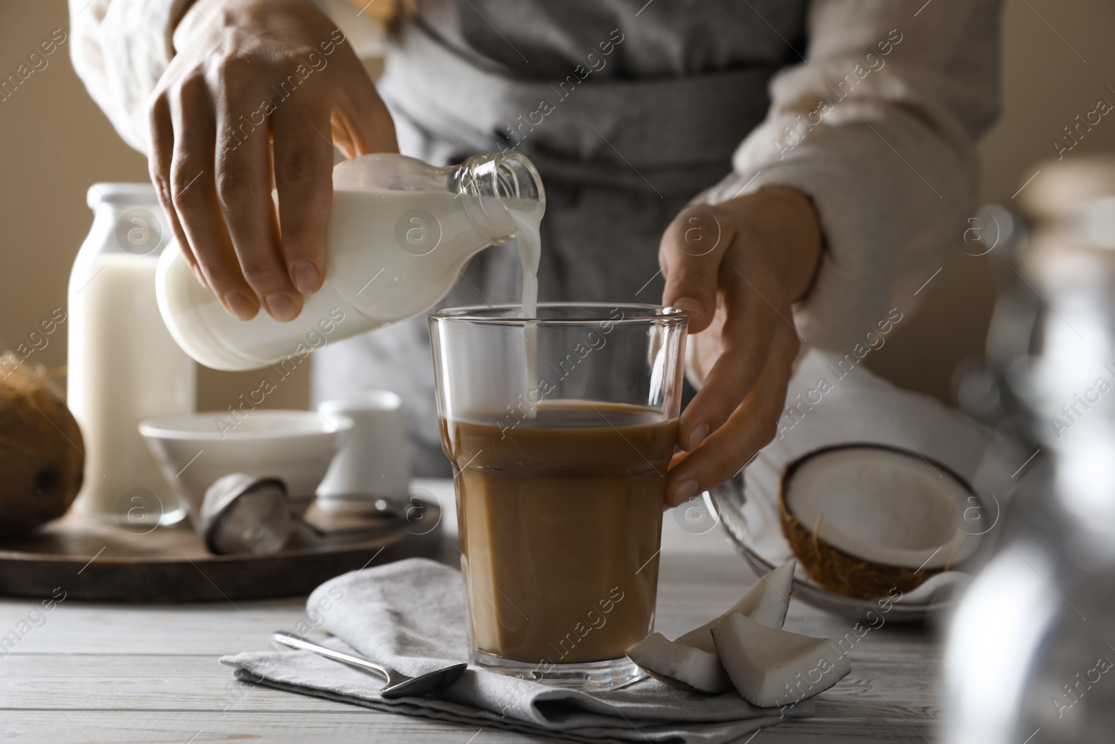 Photo of Woman pouring coconut milk into glass of coffee at white wooden table, closeup