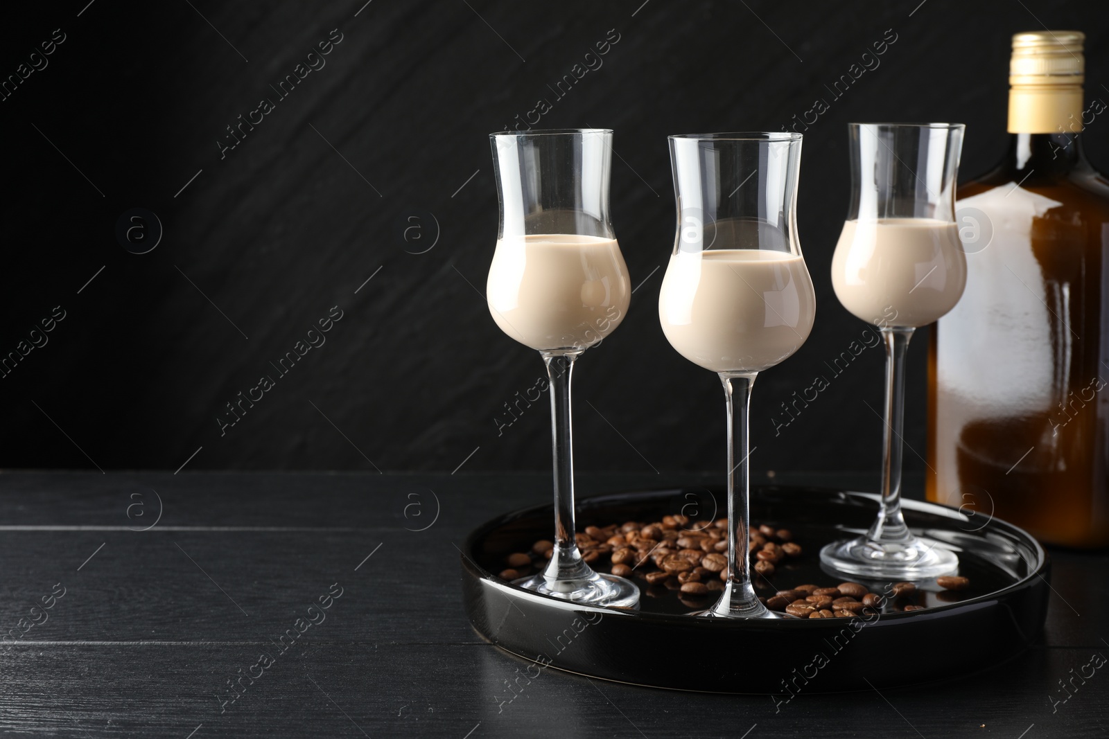 Photo of Coffee cream liqueur in glasses, bottle and beans on black wooden table, space for text