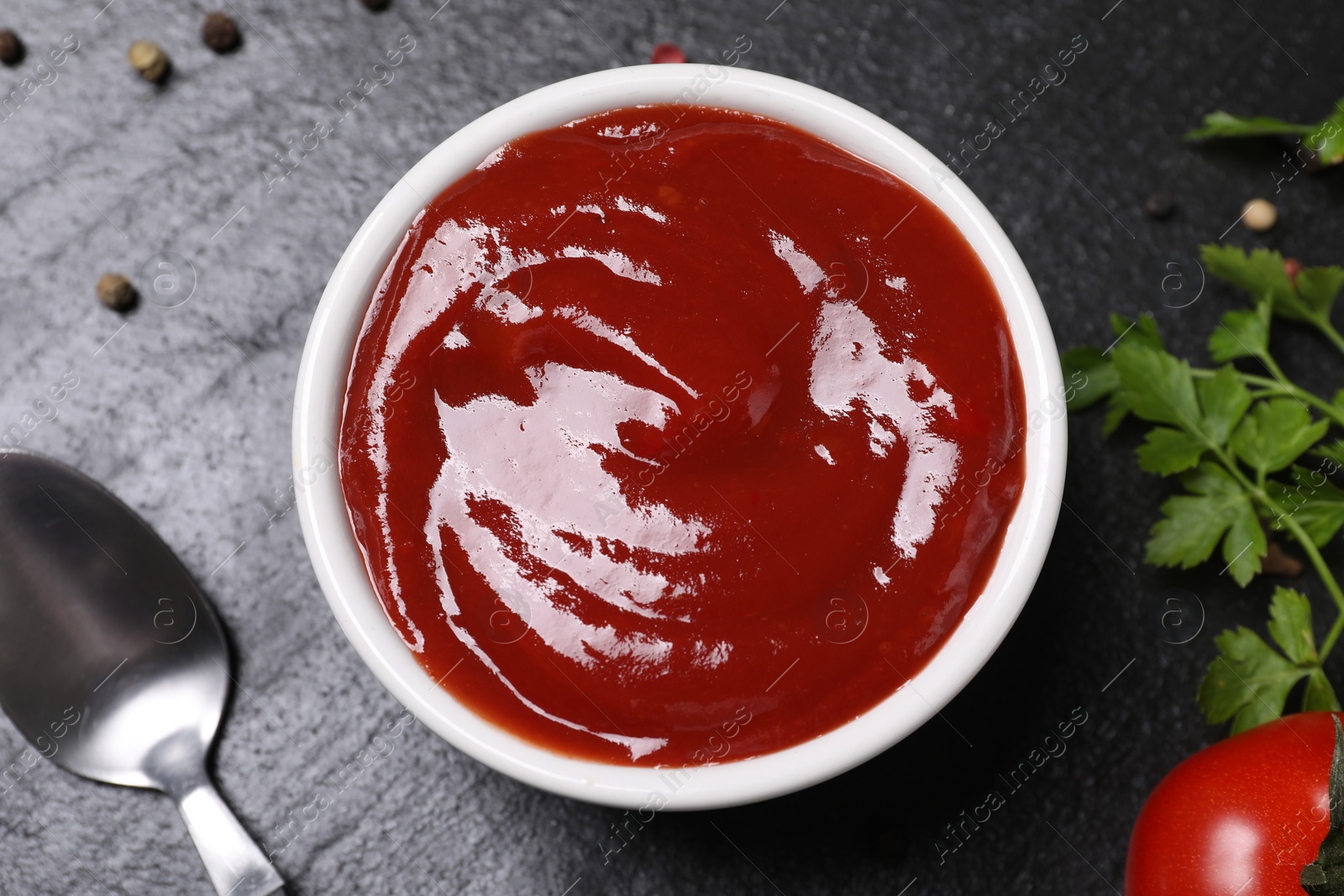 Photo of Organic ketchup in bowl and spoon on black table, flat lay. Tomato sauce