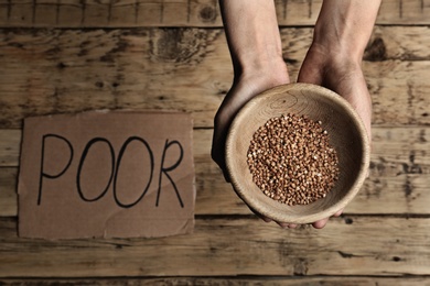 Poor woman holding bowl with grains against wooden background, closeup