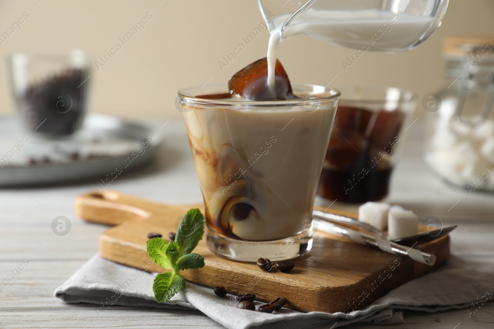 Photo of Pouring milk from jug into glass of delicious iced coffee, beans and mint on white wooden table