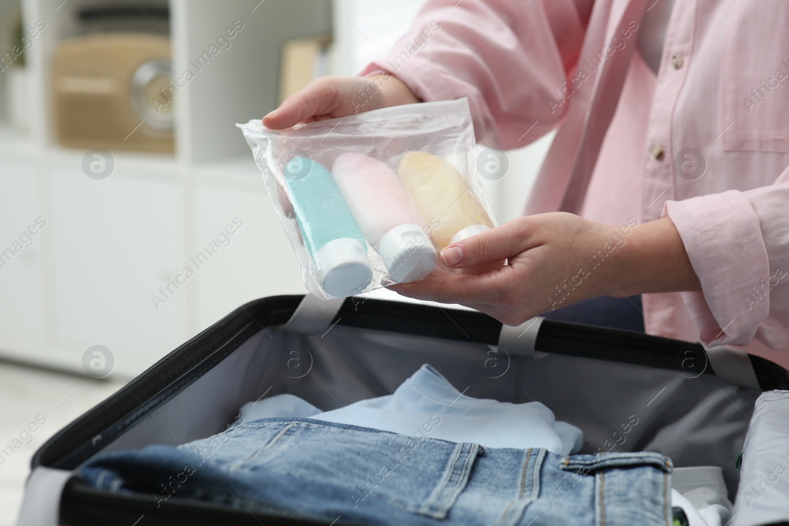 Photo of Woman holding plastic bag of cosmetic travel kit over suitcase indoors, closeup. Bath accessories