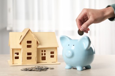 Photo of House model and money on wooden table. Woman putting coin into piggy bank indoors, closeup