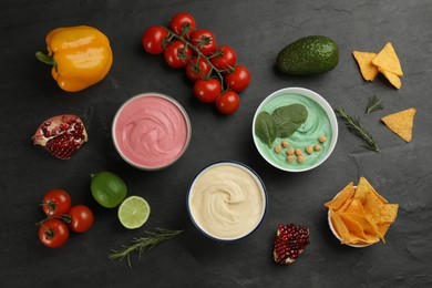 Different kinds of tasty hummus, nachos and ingredients on black table, flat lay