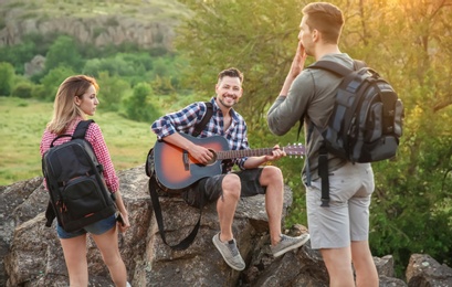 Young man with backpack playing guitar for his friends in wilderness. Camping season