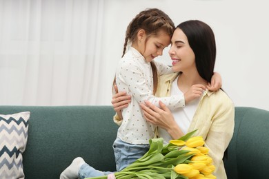Photo of Happy woman with her daughter and bouquet of yellow tulips on sofa at home, space for text. Mother's day celebration