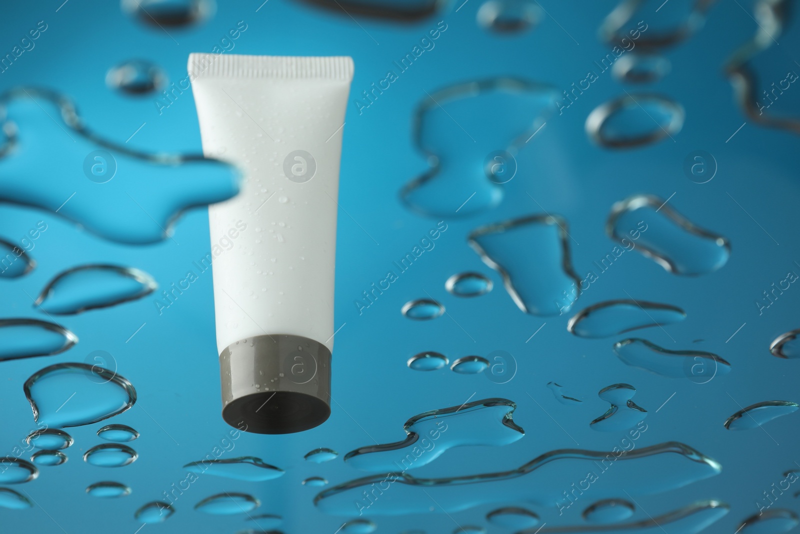 Photo of Moisturizing cream in tube on glass with water drops against blue background, low angle view. Space for text