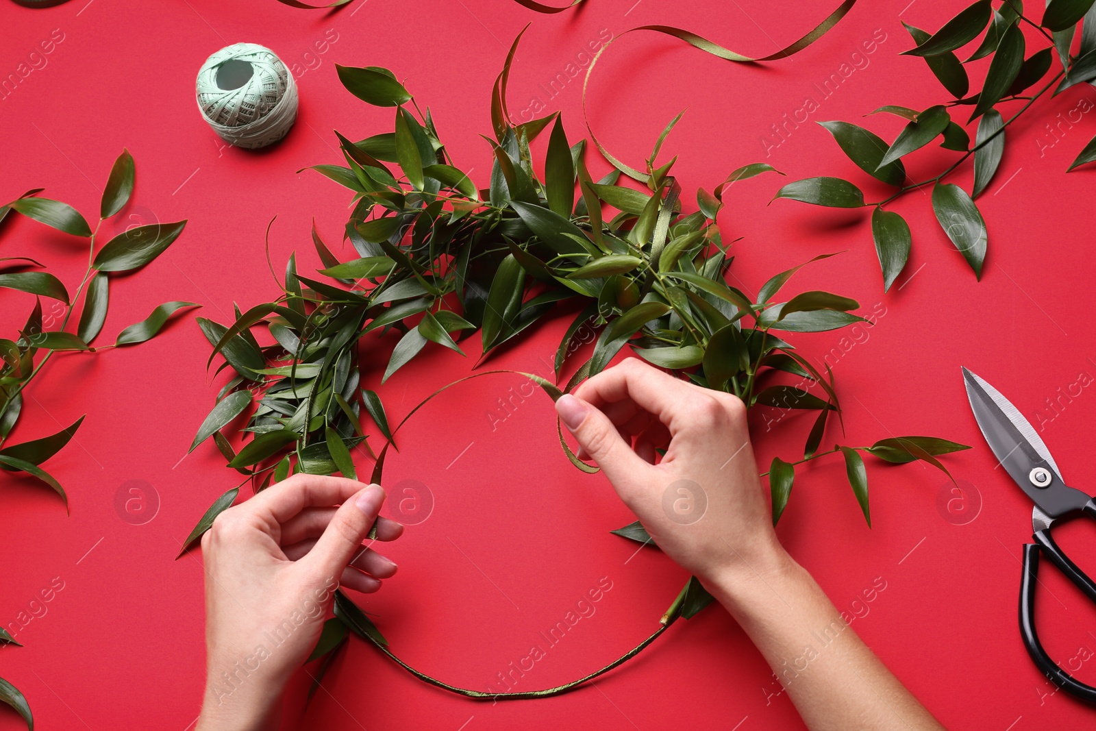 Photo of Florist making beautiful mistletoe wreath on red background, top view. Traditional Christmas decor