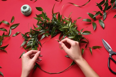 Photo of Florist making beautiful mistletoe wreath on red background, top view. Traditional Christmas decor