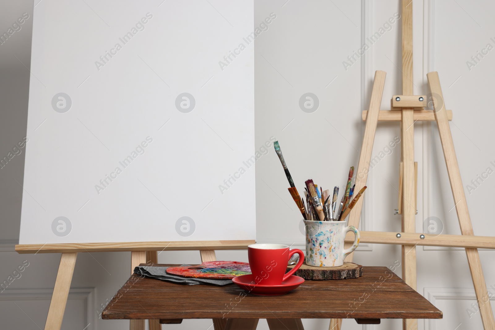 Photo of Easel with blank canvas, cup of drink and different art supplies on wooden table near white wall