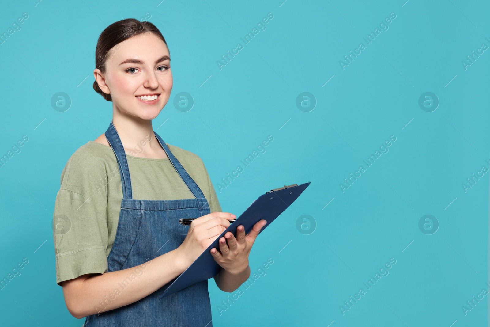 Photo of Beautiful young woman in clean denim apron with clipboard on light blue background. Space for text