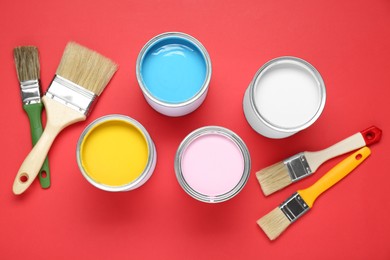 Cans of colorful paints and brushes on red background, flat lay